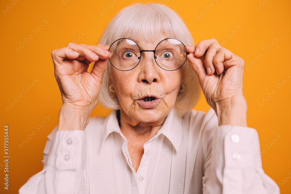 Closeup portrait of elderly surprised woman with white hair and gray eyes, wearing light blouse, adjusting round glasses and standing over bright orange background.