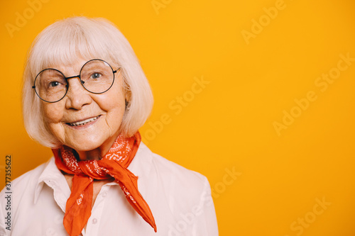 Elderly gray-haired woman with gray eyes wearing white blouse and orange scarf or cravat. Woman standing isolated over orange background, looking sideway and smiling.
