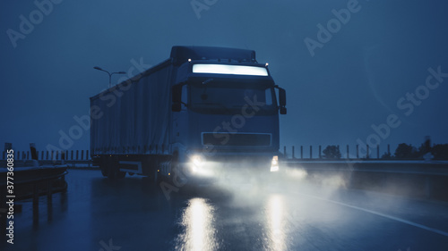 Blue Long Haul Semi-Truck with Cargo Trailer Full of Goods Travels At Night on the Freeway Road  Driving Across Continent Through Rain  Fog  Snow. Industrial Warehouses Area. Front Shot