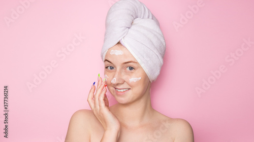 Smiling woman with a towel on her head on a pink background and face cream. Hydration of the skin, skin care face, beauty.