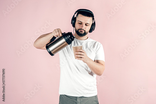 A young man in big headphones holds a paper cup in his hands, pours from a thermos into a paper cup.