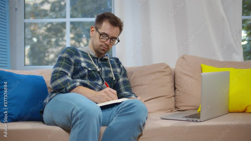 Young man having video conference on laptop writing down notes sitting on couch at home Young man having video conference on laptop writing down notes sitting on couch at home