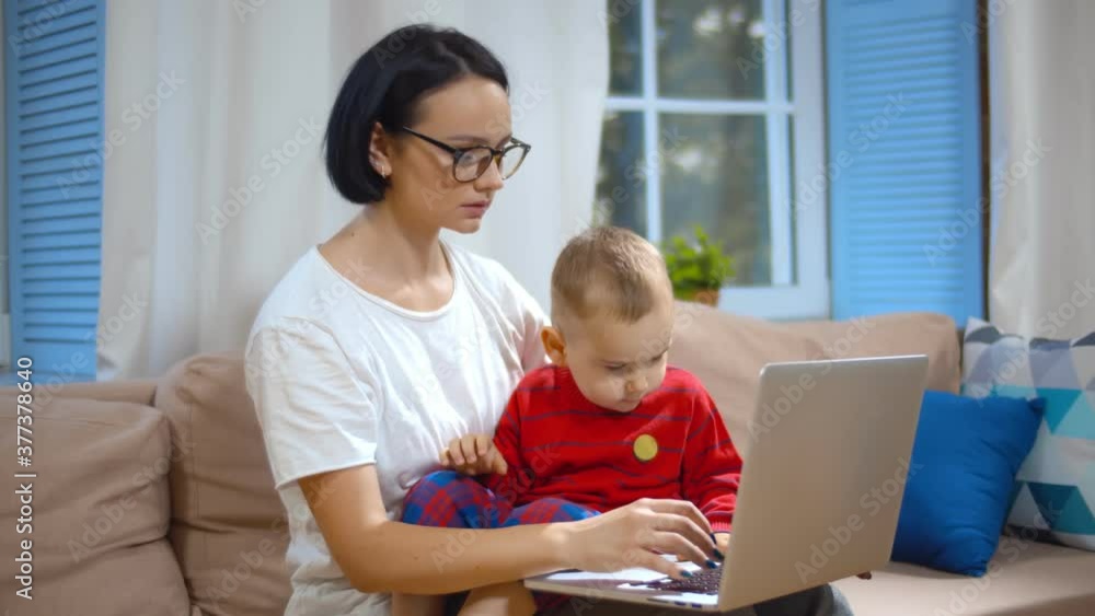 Young woman sitting on couch with baby checking financial report on laptop working from home Young woman sitting on couch with baby checking financial report on laptop working from home
