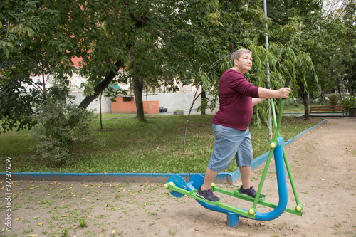 Elderly woman is doing exercise on a sports trainer ellipse. Workout in the yard. The concept of a happy old age and body positive. International Day of Older Persons and Grandparents Day