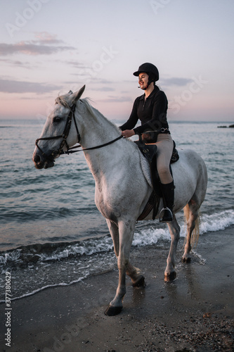 white horse with a woman riding stands on the beach in the evening