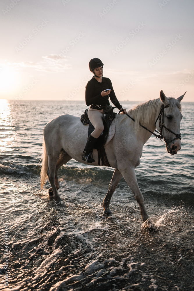 Fototapeta premium young woman with phone rides astride a white beautiful horse on the beach at sunset
