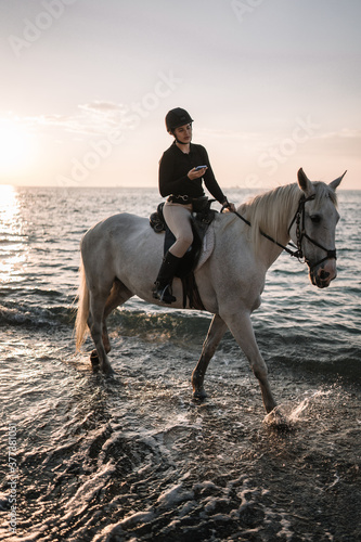 young woman with phone rides astride a white beautiful horse on the beach at sunset