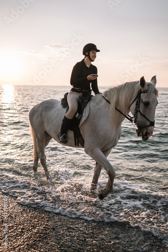 young woman with a phone rides a white beautiful horse and laughs on the beach at sunset