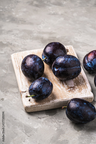 Fresh ripe plum fruits with water drops on wooden cutting board, stone concrete background, angle view