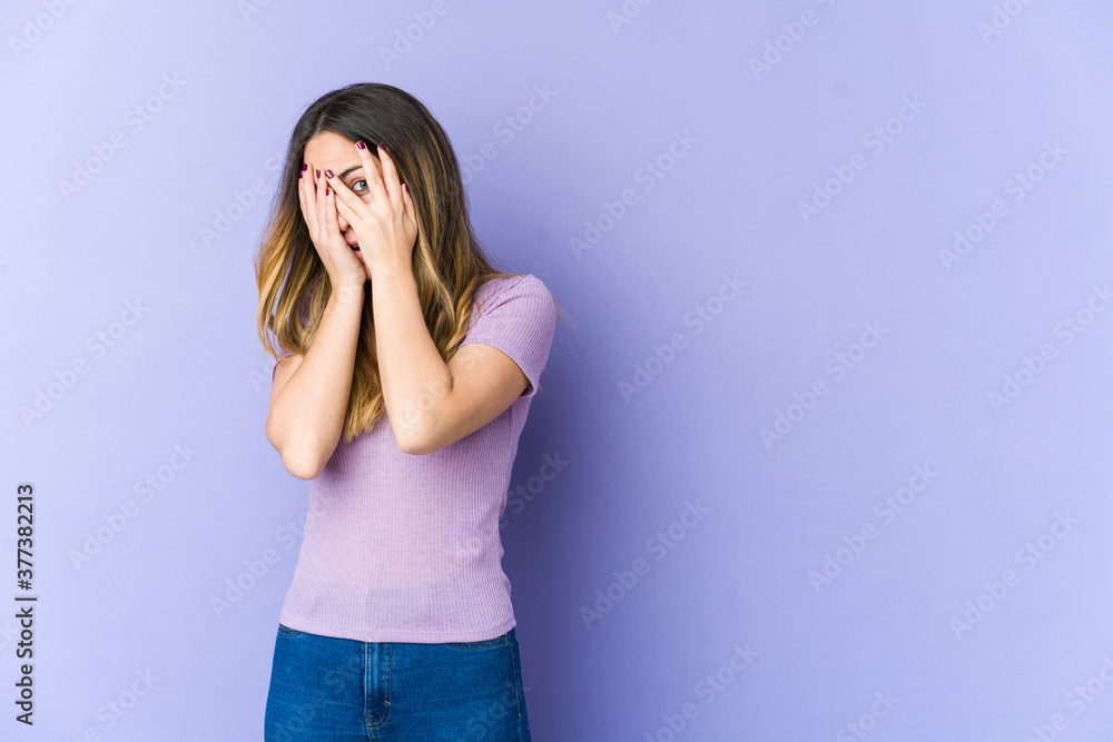 Young caucasian woman isolated on purple background blink through fingers frightened and nervous.