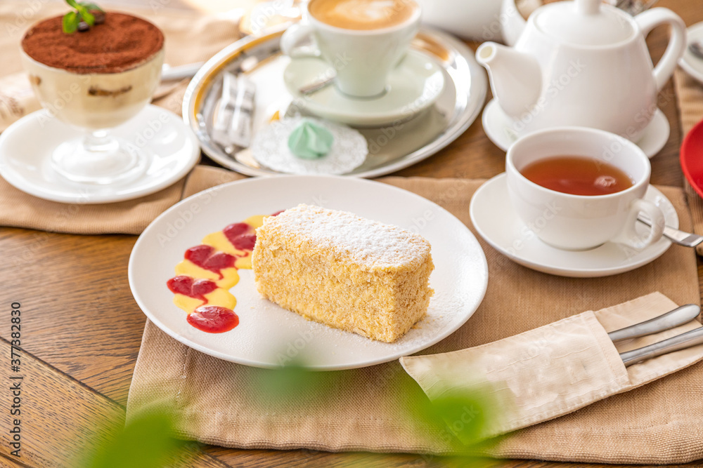 a slice of napoleon cake on a white plate on a served table. In the background there is a kettle, tea, juice.