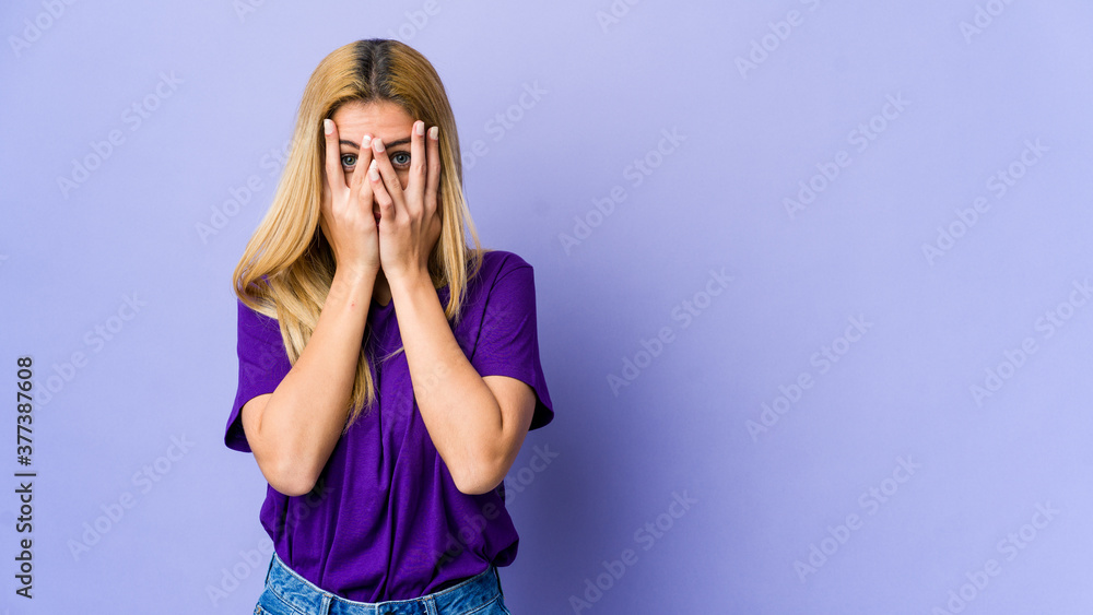 Young blonde woman isolated on purple background blink at the camera through fingers, embarrassed covering face. Young blonde woman isolated on purple background blink at the camera through fingers, embarrassed covering face.