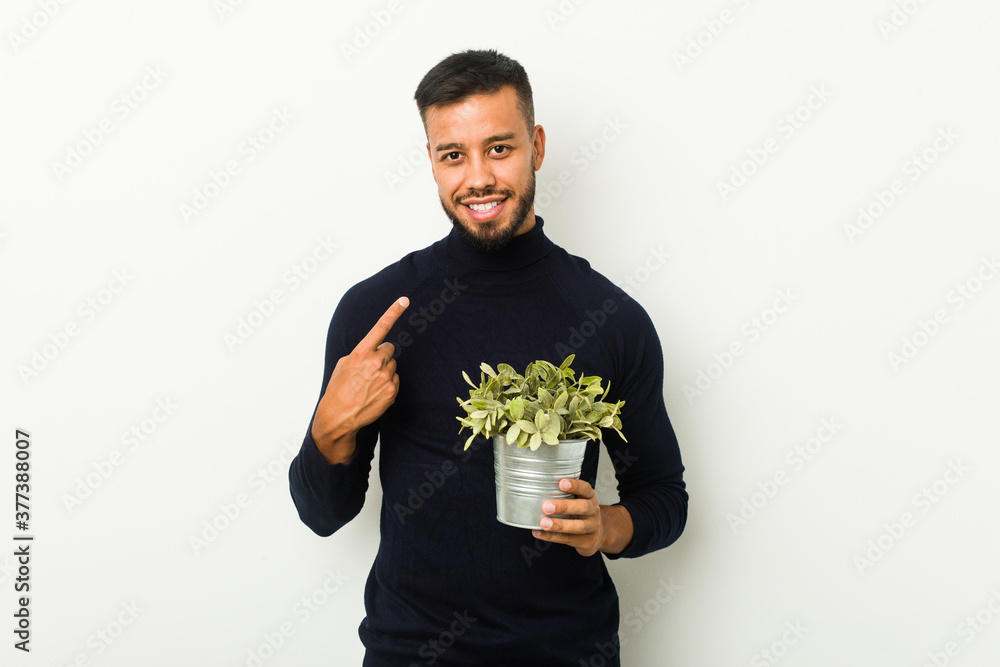 Young south-asian man holding a plant pointing with finger at you as if inviting come closer. Young south-asian man holding a plant pointing with finger at you as if inviting come closer.