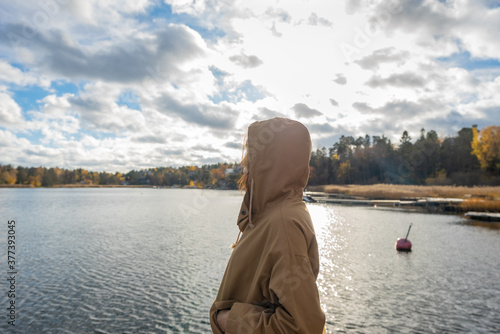 Young woman in a jacket in the fall in the rays against the setting sun. Sun glare. A girl stands on the seashore and looks at the horizon..