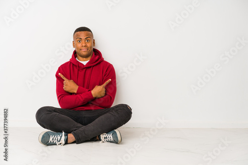 Young latin man sitting on the floor isolated points sideways, is trying to choose between two options.