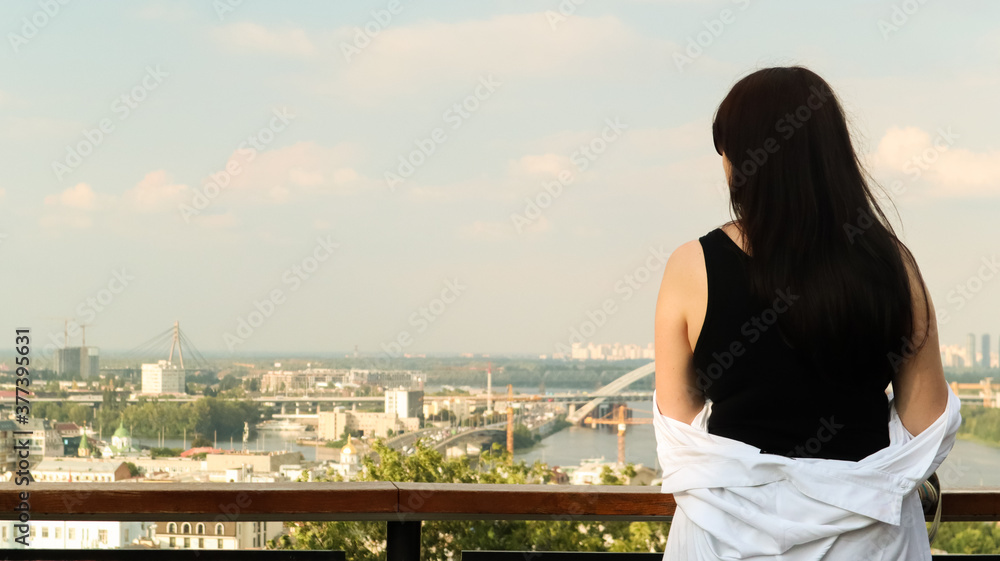 Back view of a young woman in a black T-shirt and white shirt, a brunette traveler looks at the cityscape on a summer sunny day from a high hill with an observation deck for tourists. Vintage