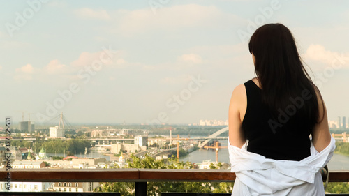 Fototapeta Naklejka Na Ścianę i Meble -  Back view of a young woman in a black T-shirt and white shirt, a brunette traveler looks at the cityscape on a summer sunny day from a high hill with an observation deck for tourists. Vintage