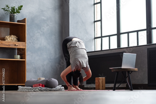 Young woman working out indoors standing in Uttanasana during online lesson.