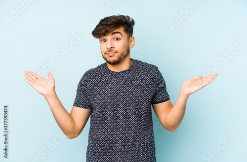 Young arabian man isolated on a blue background doubting and shrugging shoulders in questioning gesture. photo