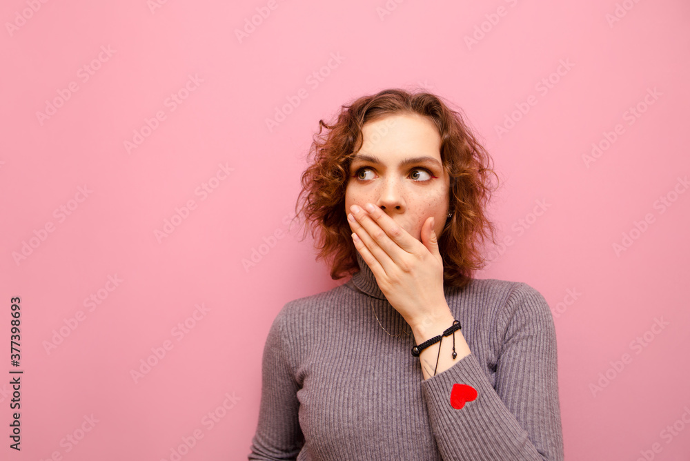 Closeup portrait of shocked attractive girl with curly hair covering mouth with her hand and looking away with frightened face isolated on pink background. Copy space Closeup portrait of shocked attractive girl with curly hair covering mouth with her hand and looking away with frightened face isolated on pink background. Copy space