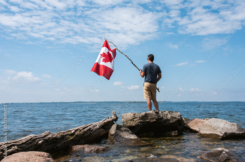 Man holding Canada flag on rocky shore of a lake on a summer day.