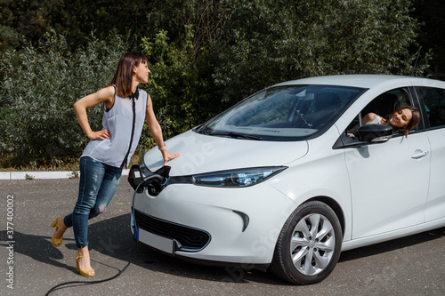 Two happy young beautiful women are talking to each other until their electric car is charging at the charging station situated in the forest. © Med Photo Studio