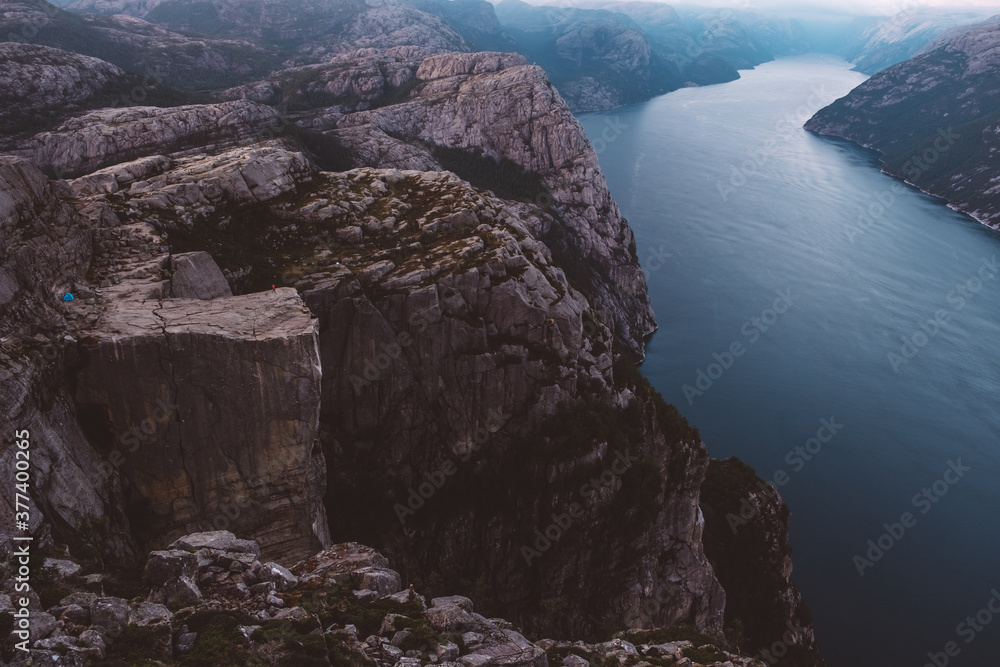 View of Norwegian fjords in cloudy day, blue camping tent and unrecognizable person on red jacket standing on cliff. View of Norwegian fjords in cloudy day, blue camping tent and unrecognizable person on red jacket standing on cliff.