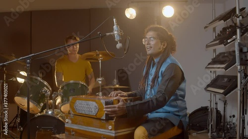Music recording. Indian man playing harmonium in the recording studio with caucasian man on drums. Hindi male person in national clothes plays meditation music. photo