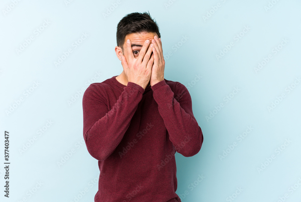 Young caucasian man isolated on blue background blink through fingers frightened and nervous. Young caucasian man isolated on blue background blink through fingers frightened and nervous.
