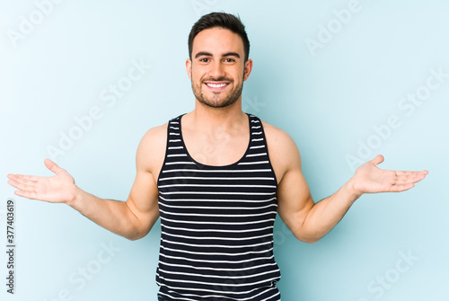 Young caucasian man isolated on blue background makes scale with arms, feels happy and confident.