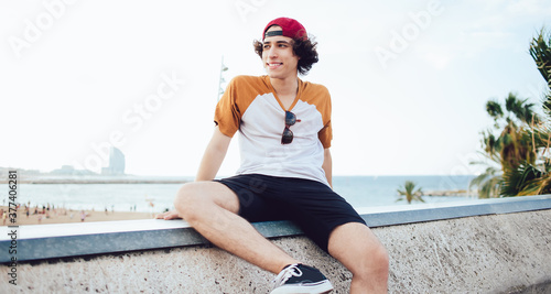 Carefree teenager 20 years old resting after promenade on weekend sitting at urbanity and smiling, happy Caucasian man dressed in casual clothing recreating near seashore enjoying summer vacations #377406281