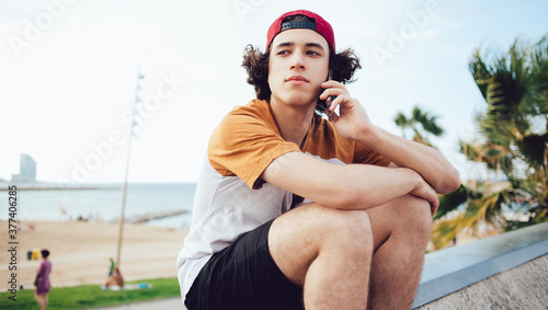 Fototapeta Naklejka Na Ścianę i Meble -  Millennial male teenager in red cap making international cellphone communication sitting at urbanity, youthful hipster guy calling via smartphone application connecting to roaming internet