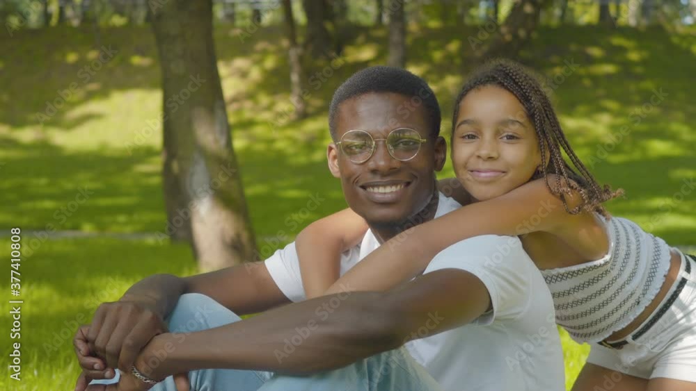 Side view of cute girl hugging man in sunny summer park. Portrait of happy African American sister and brother looking at camera and smiling. Joy and leisure concept. Side view of cute girl hugging man in sunny summer park. Portrait of happy African American sister and brother looking at camera and smiling. Joy and leisure concept.