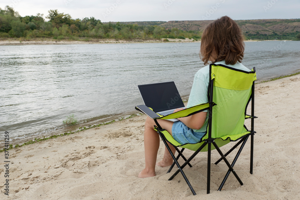 young female with laptop in her knees is sitting camping chair on the bank of picturesque river. Frelancer and remote work concept. Opportunity for distant work.