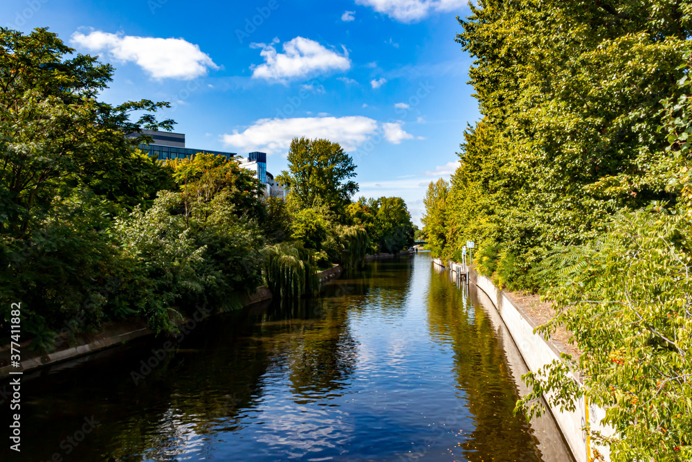 A sunny day in Berlin, Germany. Image taken from a bridge in the city center features one of many canals inside the city with trees on both sides, reflection in water and clear sky in background