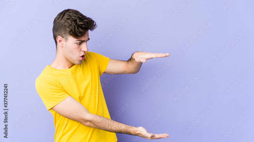 Young caucasian man isolated on purple background shocked and amazed holding a copy space between hands. Young caucasian man isolated on purple background shocked and amazed holding a copy space between hands.