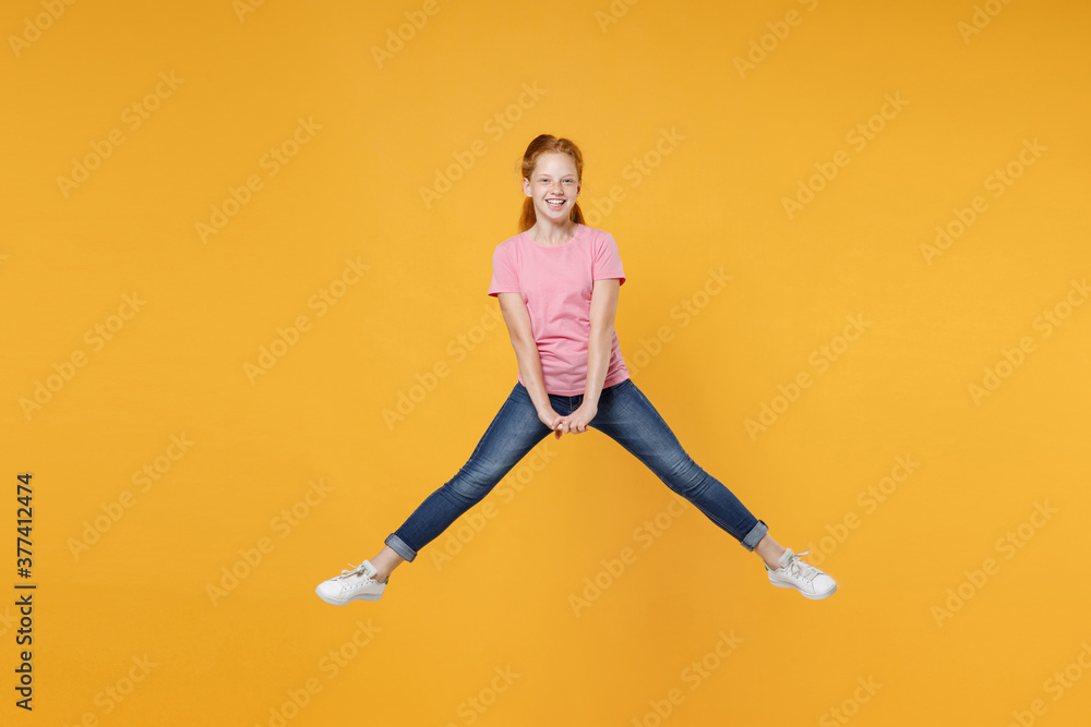 Full length children studio portrait of smiling little ginger redhead kid girl 12-13 years old wearing pink casual t-shirt posing jumping spreading legs isolated on bright yellow color background.