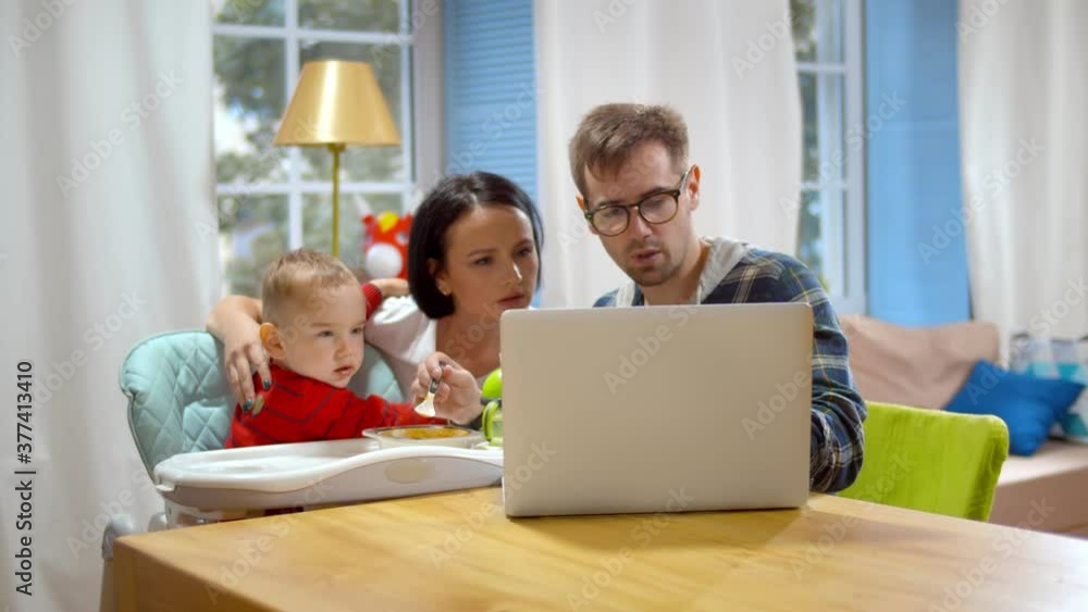 Happy young couple feeding baby boy in highchair and using laptop at home Happy young couple feeding baby boy in highchair and using laptop at home