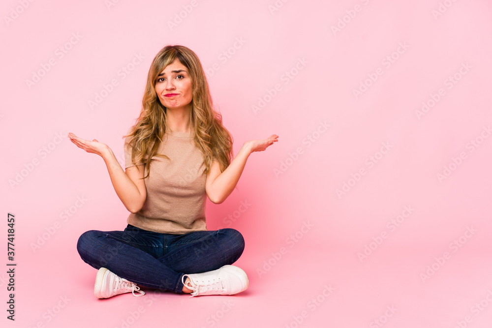 Young blonde caucasian woman sitting on a pink studio doubting and shrugging shoulders in questioning gesture. Young blonde caucasian woman sitting on a pink studio doubting and shrugging shoulders in questioning gesture.
