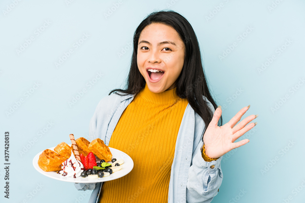 Young chinese woman holding waffle isolated on blue background pointing to the side Young chinese woman holding waffle isolated on blue background pointing to the side