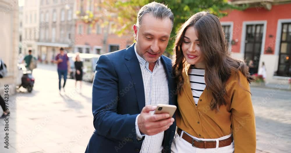 Happy Caucasian father walking with adult beautiful daughter, using smartphone and talking. Having talk. Outdoor. Cheerful man and girl strolling, watching on mobile phone and smiling. Cozy street. Happy Caucasian father walking with adult beautiful daughter, using smartphone and talking. Having talk. Outdoor. Cheerful man and girl strolling, watching on mobile phone and smiling. Cozy street.