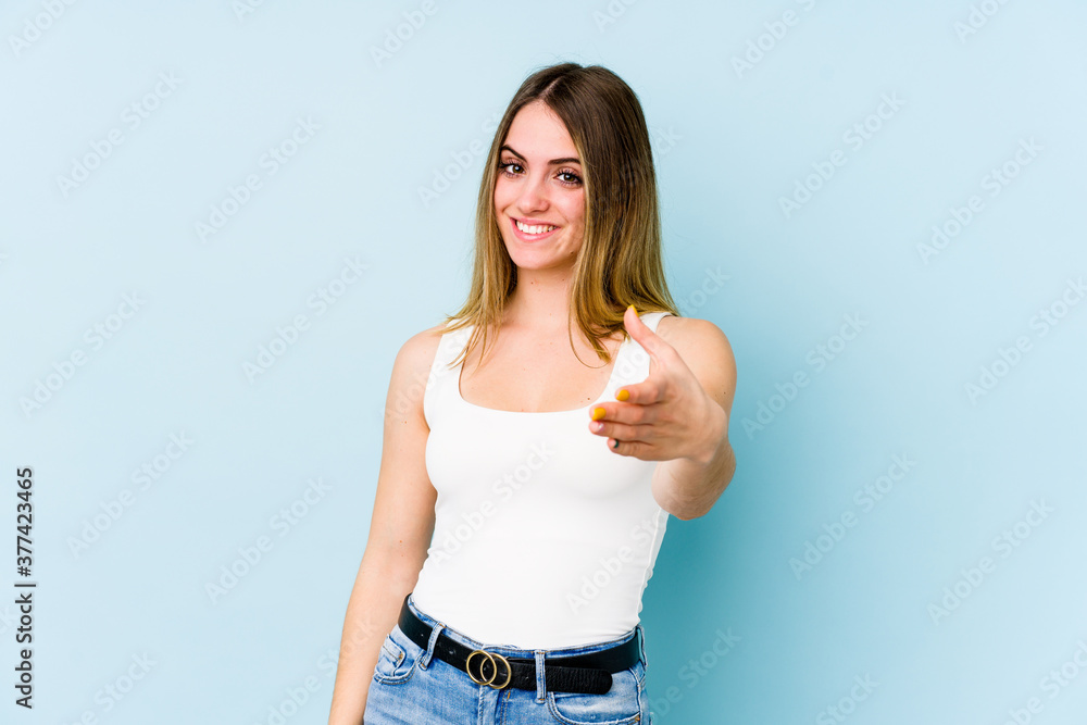 Young caucasian woman isolated on blue background stretching hand at camera in greeting gesture. Young caucasian woman isolated on blue background stretching hand at camera in greeting gesture.