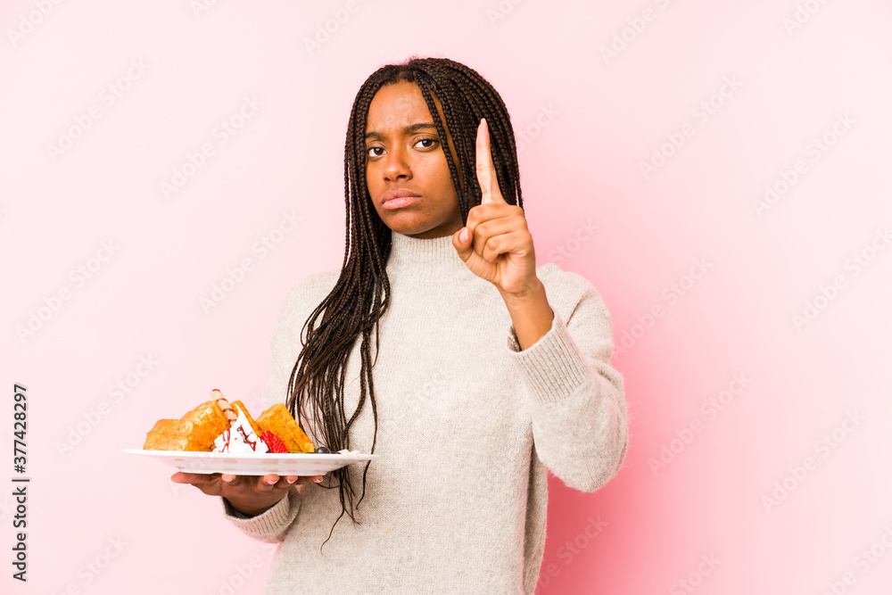 Young african american woman holding a waffle isolated showing number one with finger. Young african american woman holding a waffle isolated showing number one with finger.