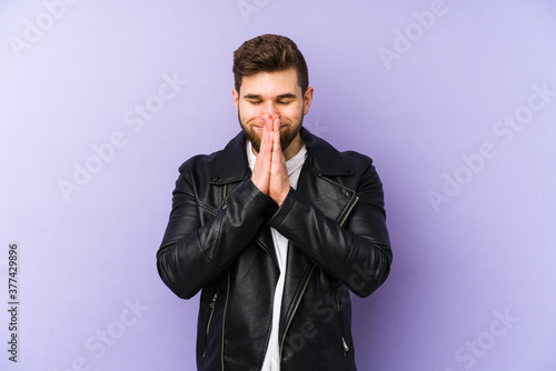 Young man isolated on purple background holding hands in pray near mouth, feels confident. © Asier