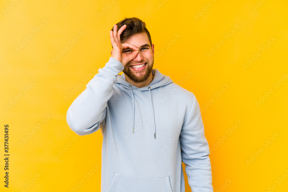 Young caucasian man isolated on yellow background excited keeping ok gesture on eye. Young caucasian man isolated on yellow background excited keeping ok gesture on eye.