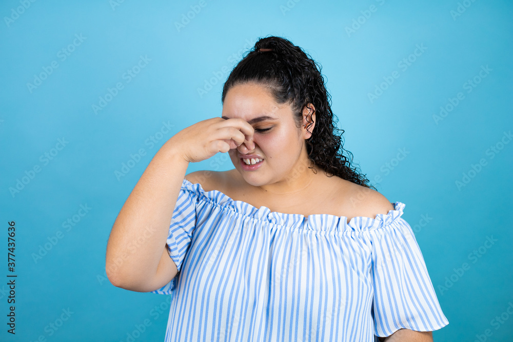 Young beautiful woman with curly hair over isolated blue background smelling something stinky and disgusting, intolerable smell, holding breath with fingers on nose Young beautiful woman with curly hair over isolated blue background smelling something stinky and disgusting, intolerable smell, holding breath with fingers on nose
