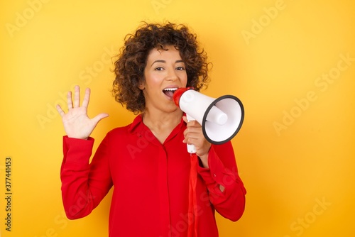 Tablou canvas Young arab woman with curly hair wearing red shirt holding a megaphone over yellow background showing and pointing up with fingers number five while smiling confident and happy