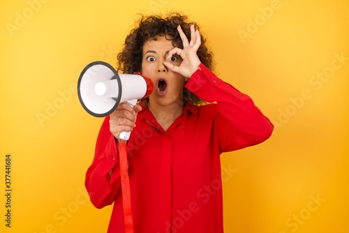 Young arab woman with curly hair wearing red shirt holding a megaphone over yellow background doing ok gesture shocked with surprised face, eye looking through fingers. Unbelieving expression.