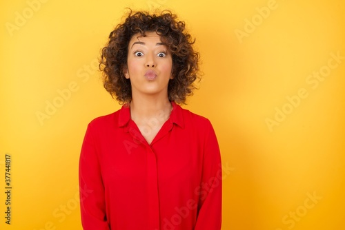 Shot of pleasant looking Young arab woman with curly hair wearing red shirt on yellow background pouts lips, looks at camera, has long hair, , has fun with girlfriend, Human facial expressions