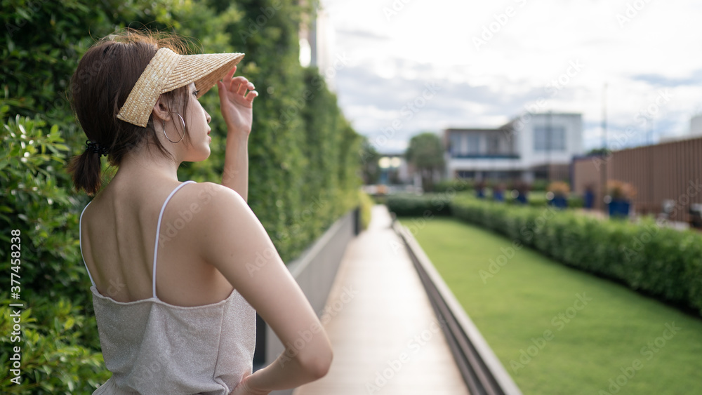 Fototapeta premium Outdoor summer portrait back view of woman with straw hat on road and green garden.